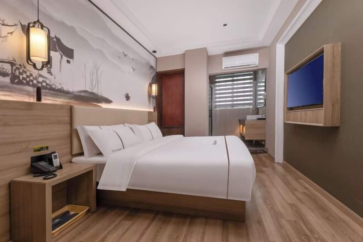 A modern hotel room with a wooden bed, a nightstand, a wall-mounted TV, and a large window with blinds.