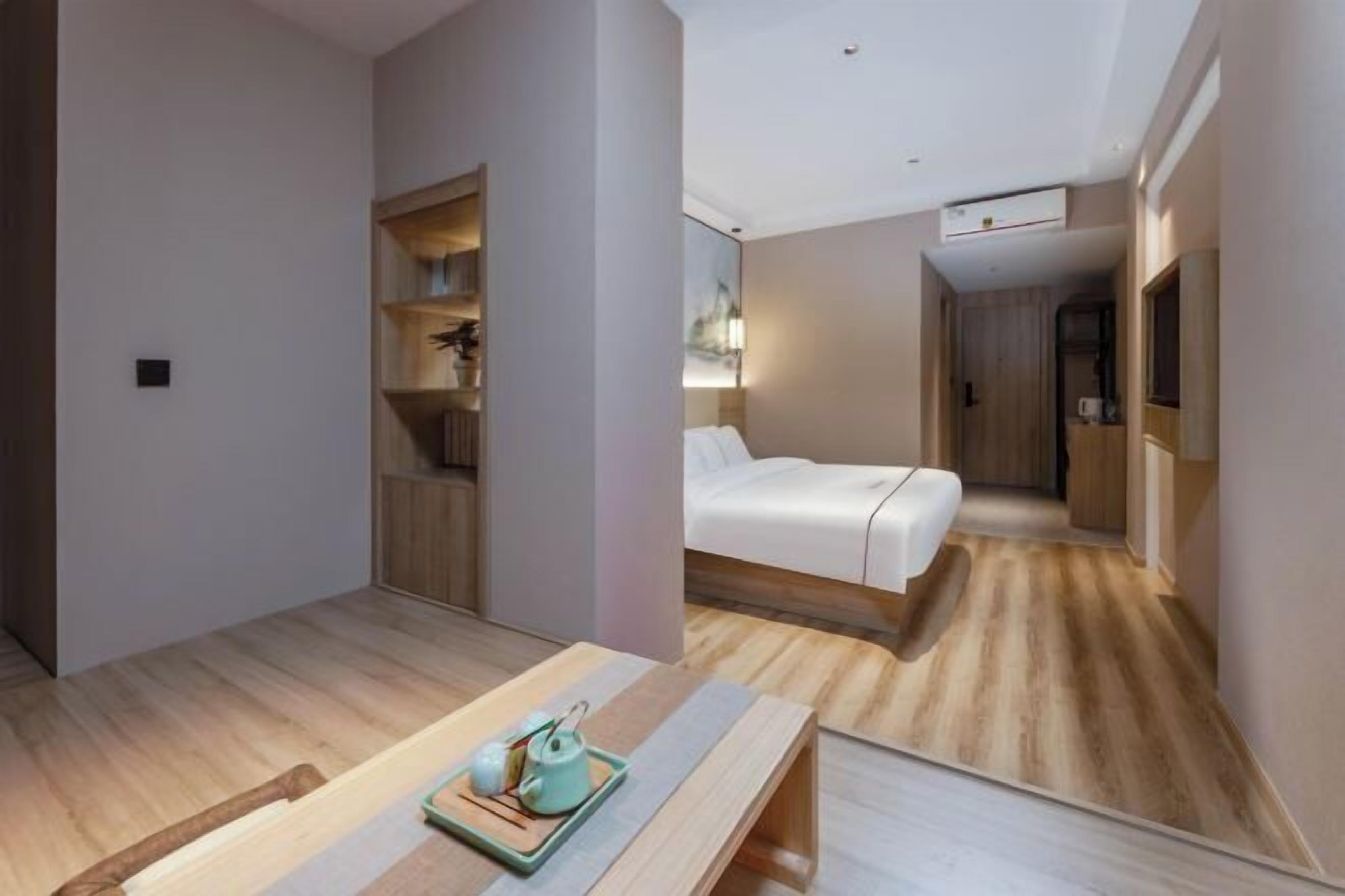 A modern hotel room with a wooden floor, a bed with white bedding, a bedside table with a teapot, and a small shelf with decorative items.