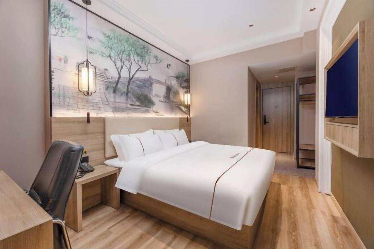 A modern hotel room with a large bed, a desk with a chair, a wardrobe, and a wall-mounted TV.