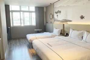 Et hotelværelse med to senge, et stort vindue, en lampe fastgjort til væggen og et lille bord.
