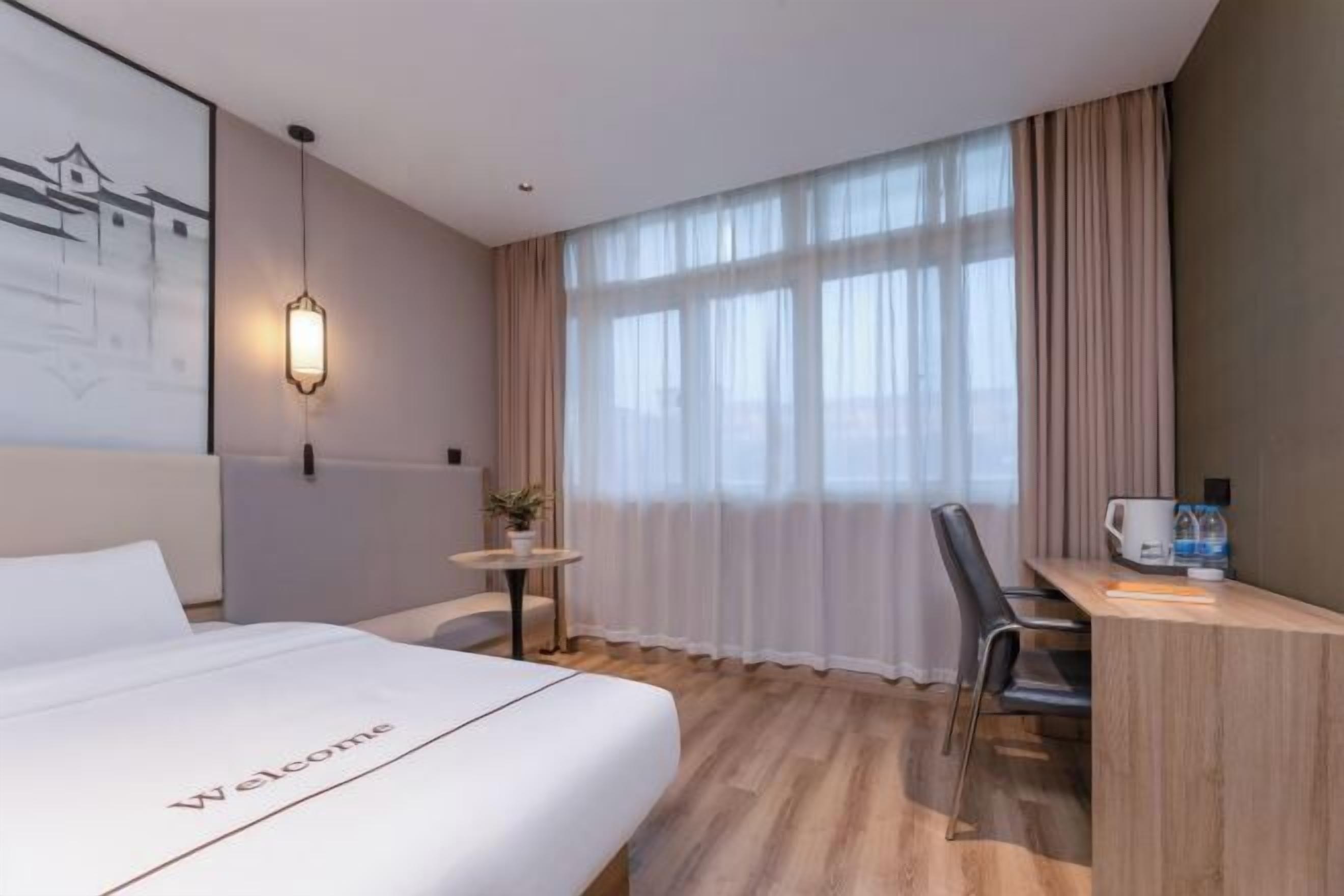 Ein Hotelzimmer mit Bett, Schreibtisch, Stuhl, einem kleinen Tisch mit Pflanze und einem Fenster mit Vorhängen.