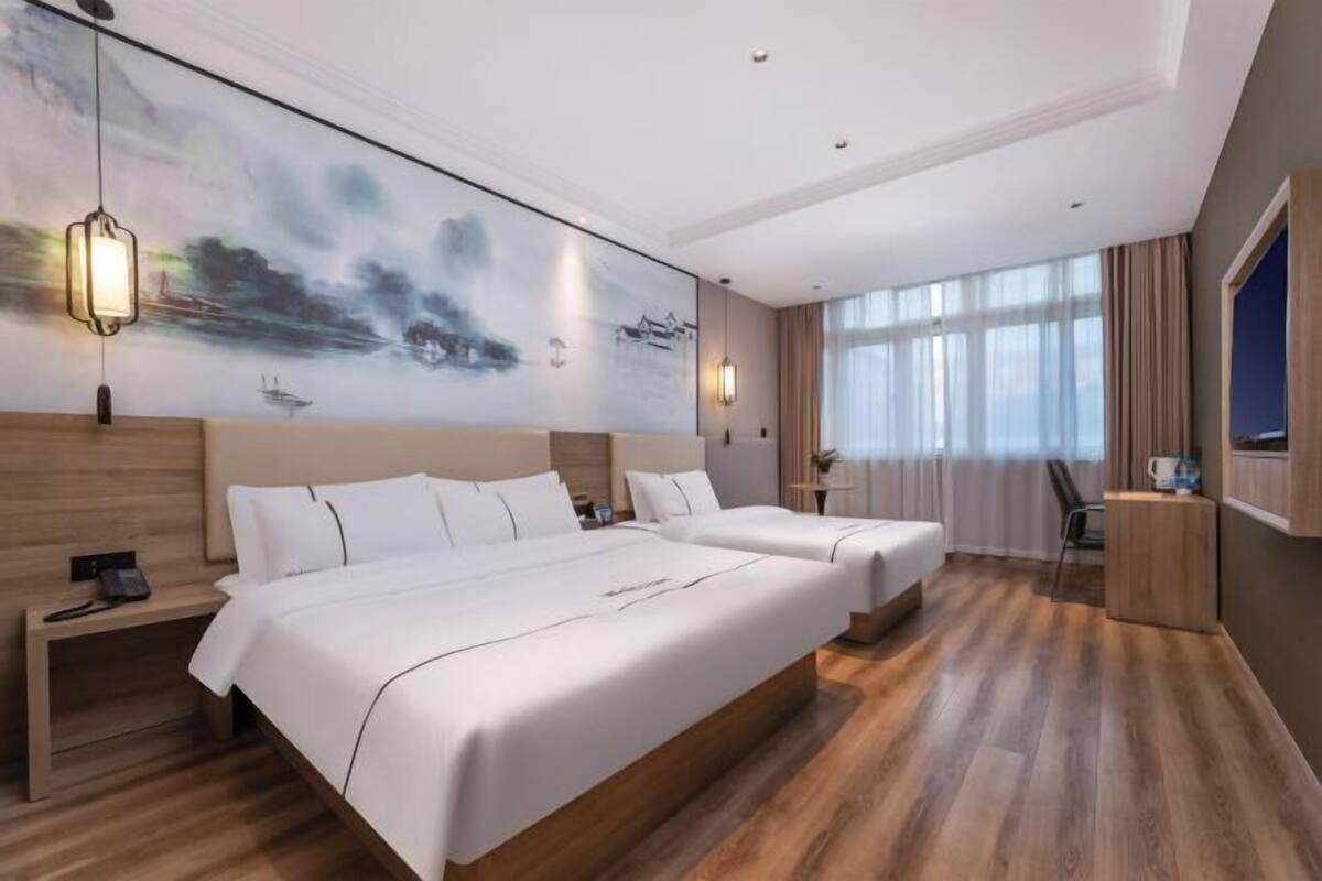 A hotel room with two beds, a large painting on the wall, a wooden floor, and a window with curtains.