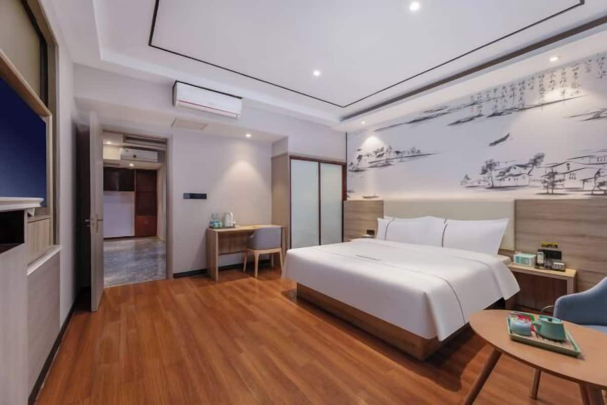 A hotel room with a bed, bedside table, chair, and a desk with a chair. There is a wall with a painting and a ceiling with recessed lighting.