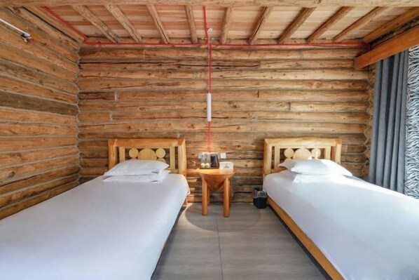 Una habitación estilo cabaña de troncos con dos camas individuales, una pequeña mesa de madera y una tubería roja que atraviesa el techo.