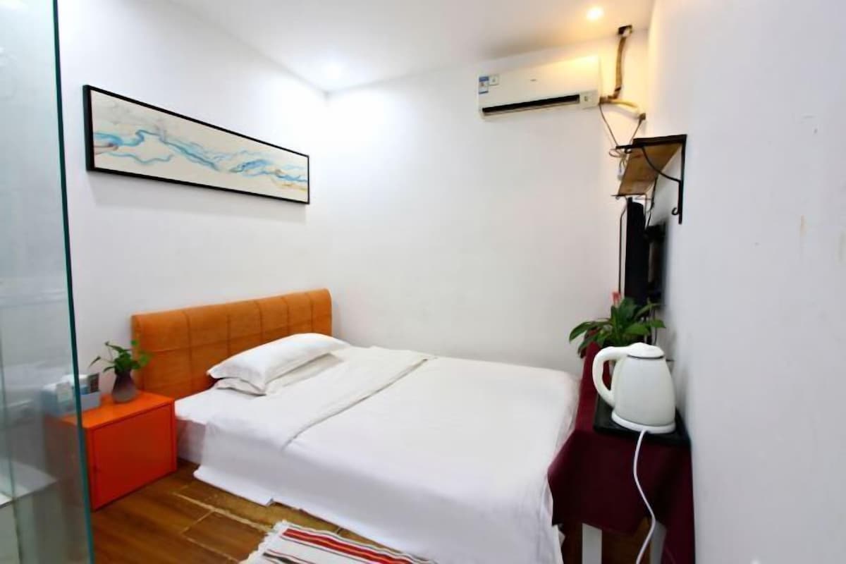 A bedroom with a bed, a bedside table with a plant, a wall-mounted air conditioner, and a painting.