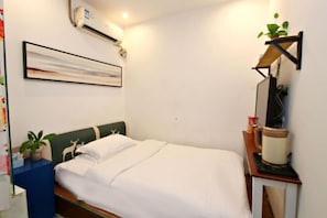 A neatly made bed with white bedding, a mounted air conditioner, a wall-mounted shelf with a plant, and a small table with a kettle. - Style Theme Hotel (Fuyang Normal University West Lake Campus) (Fuyang)