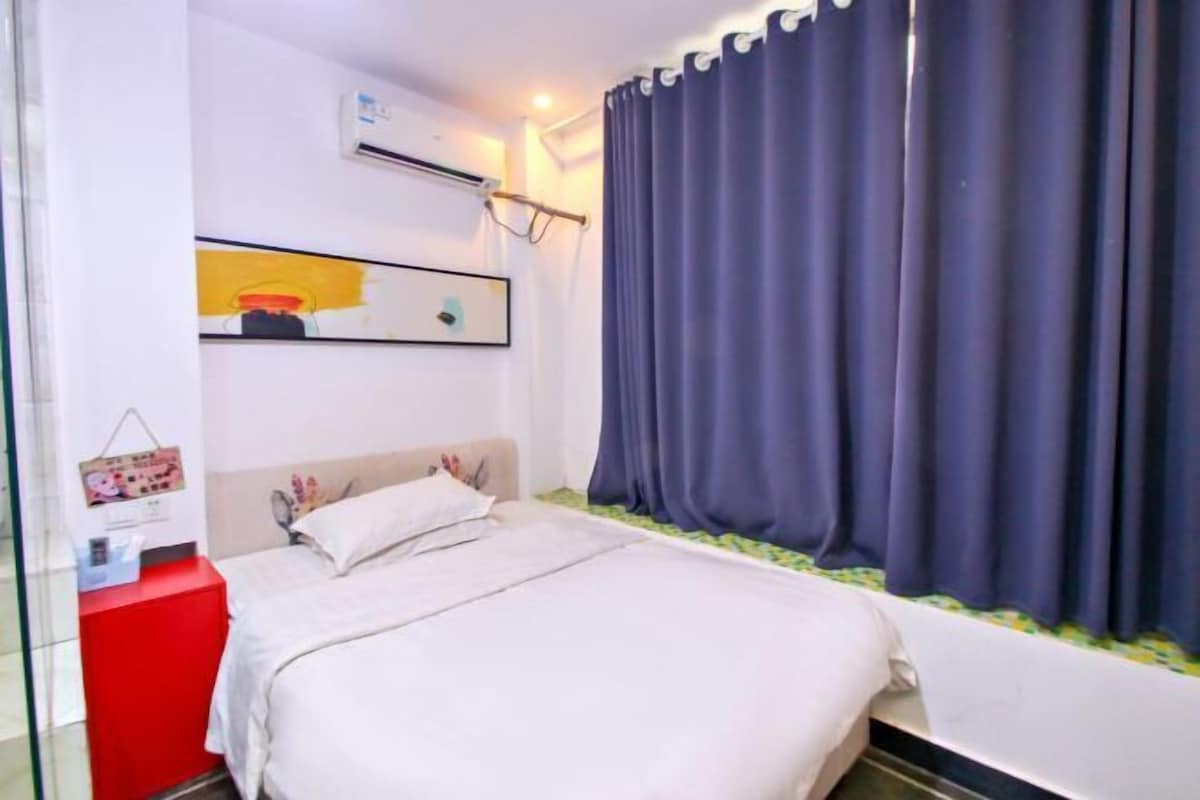 A bedroom with a bed, a red nightstand, a wall-mounted air conditioner, a window with curtains, and a framed picture.
