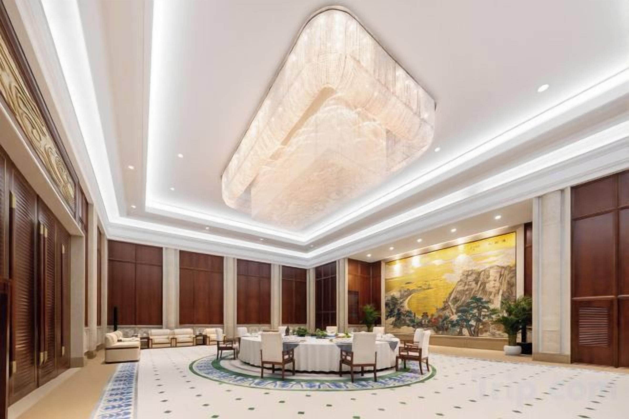 A spacious hotel lobby with a high ceiling, a large chandelier, a round dining table, and a mural on the wall.