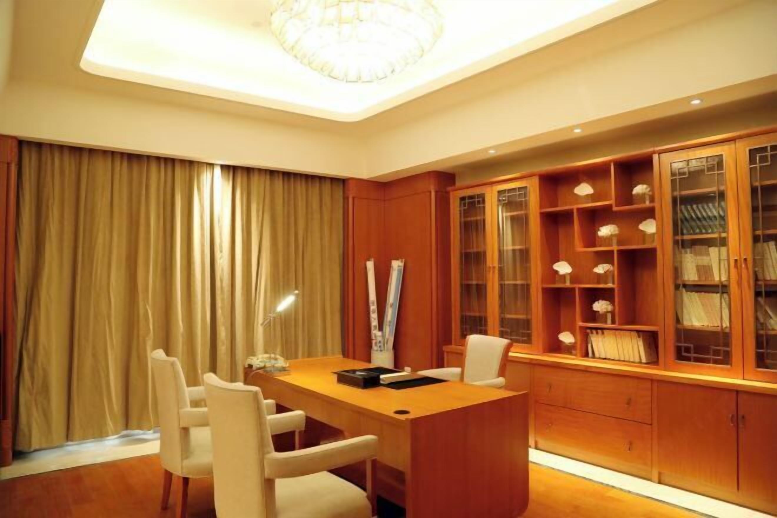 A conference room with a wooden desk, white chairs, a chandelier, and built-in shelving with books and decorative items.