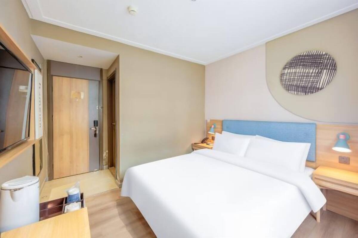 Et hotelværelse med seng, sengebord, lampe og et TV på væggen.