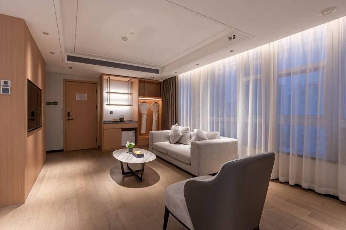 Ein modernes Hotelzimmer mit einer Couch, einem Sessel, einem kleinen Tisch, einer Küchenzeile und einem großen Fenster mit durchsichtigen Vorhängen.