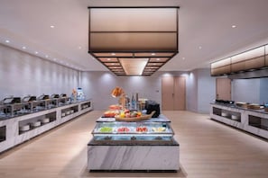 A modern hotel dining area with a central buffet, multiple food stations, and a contemporary kitchen in the background.