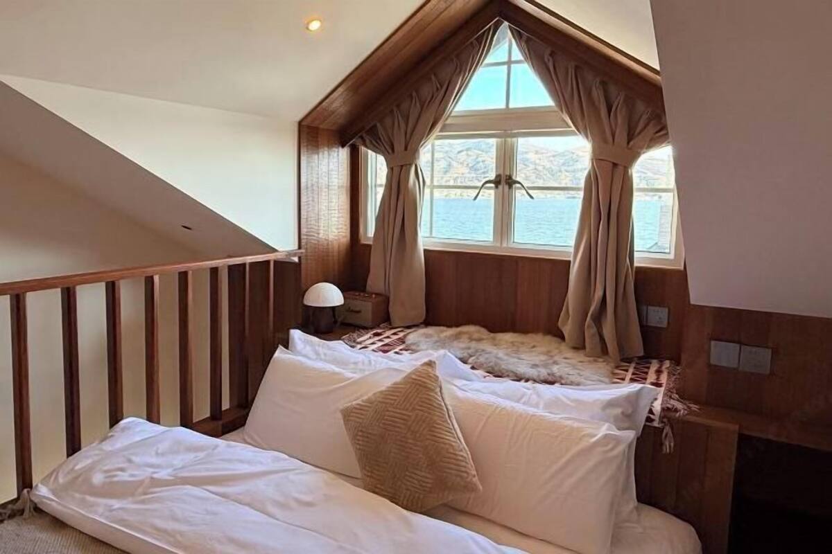A bedroom with a bed, a window with curtains, a lamp, and a view of the water.