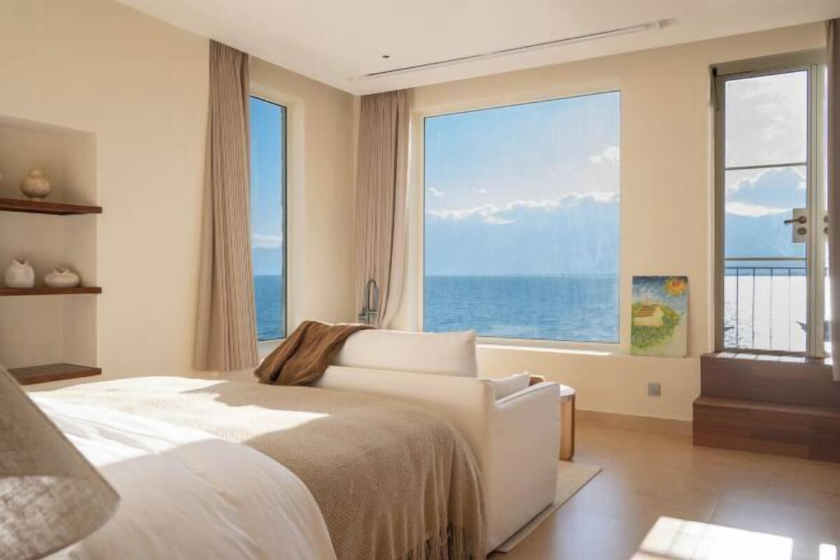 A bedroom with a large bed, a view of the sea, a window with curtains, and a painting on the wall.