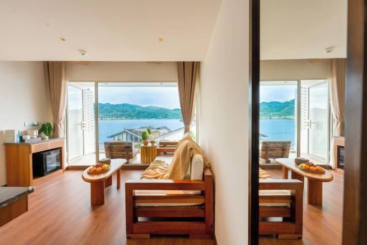 A modern room with a large window overlooking a body of water and hills. There is a wooden bench with a cushion and a small table with fruit.