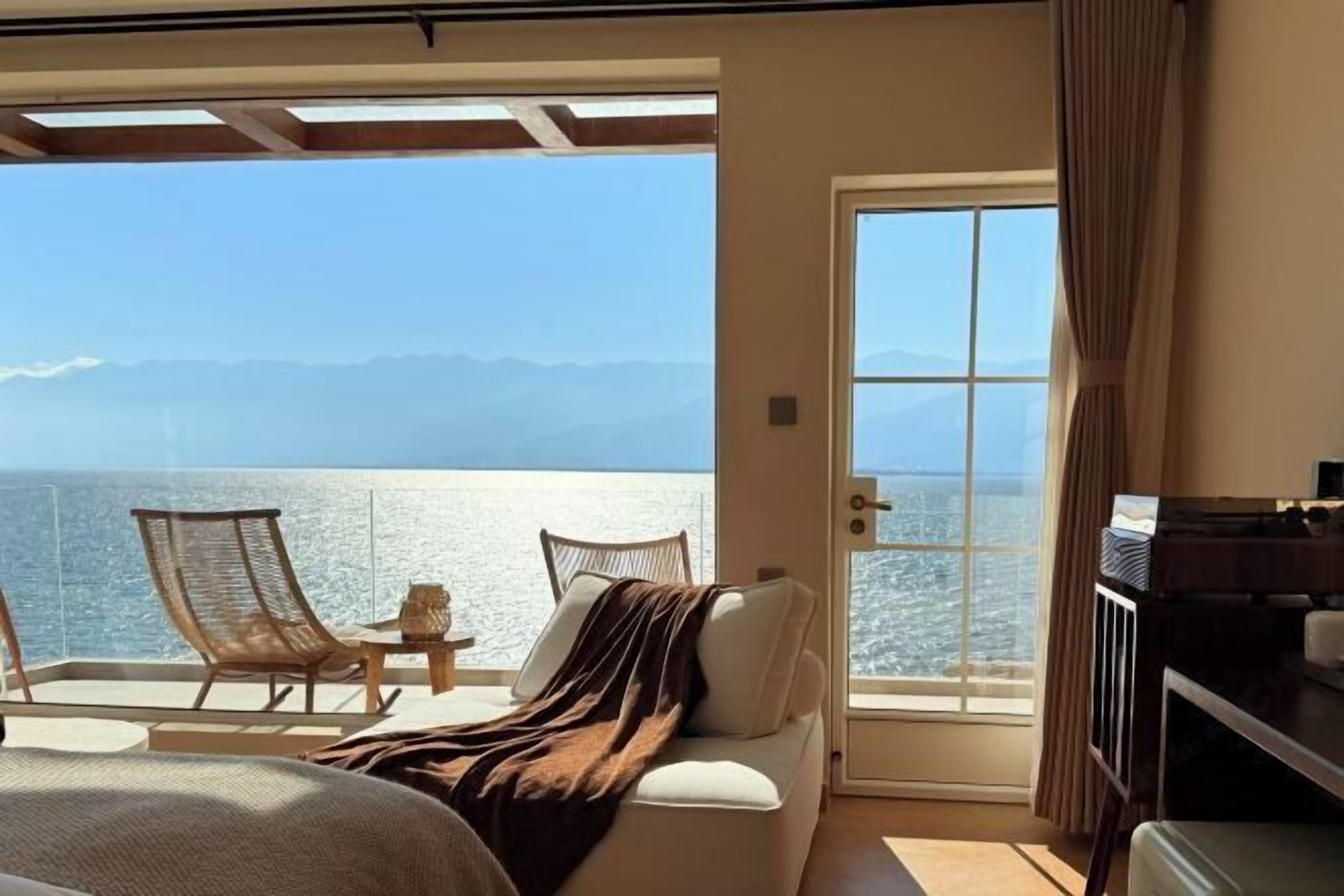 A balcony with a view of the sea and mountains, a bed, a TV, and a table with a lamp.