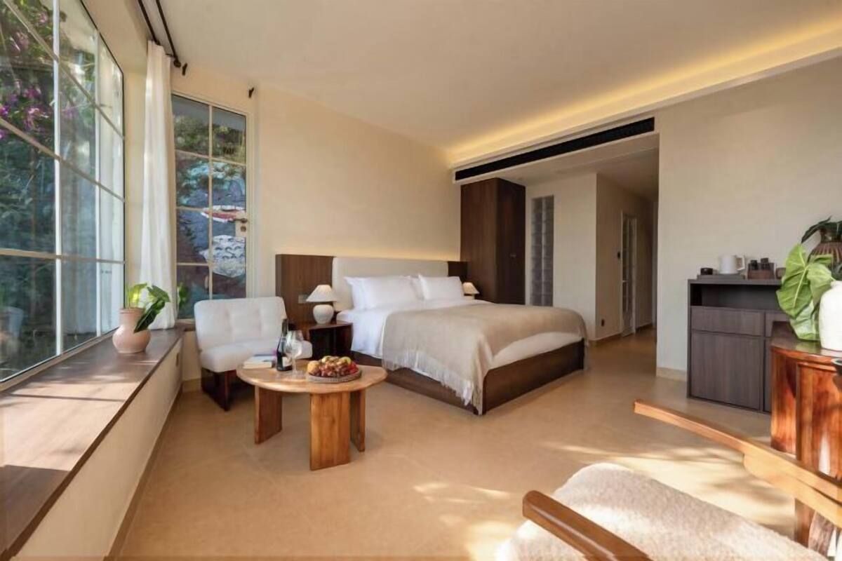 A modern hotel room with a large bed, a seating area with a table, and a view through large windows.