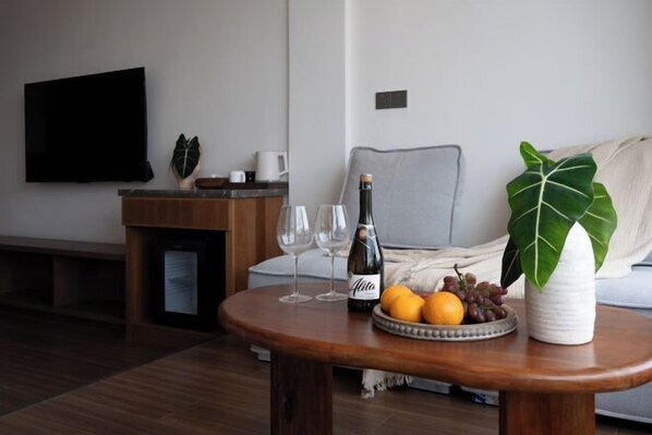 A living room with a wooden coffee table, a bottle of wine, two wine glasses, a bowl of fruit, and a potted plant.