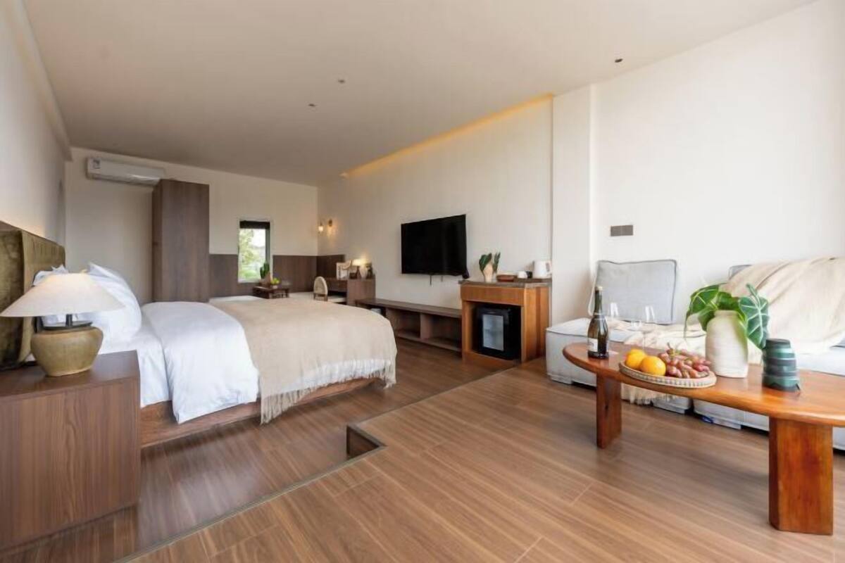 A modern hotel room with a bed, a sofa, a coffee table with fruits, a TV, and a fireplace.