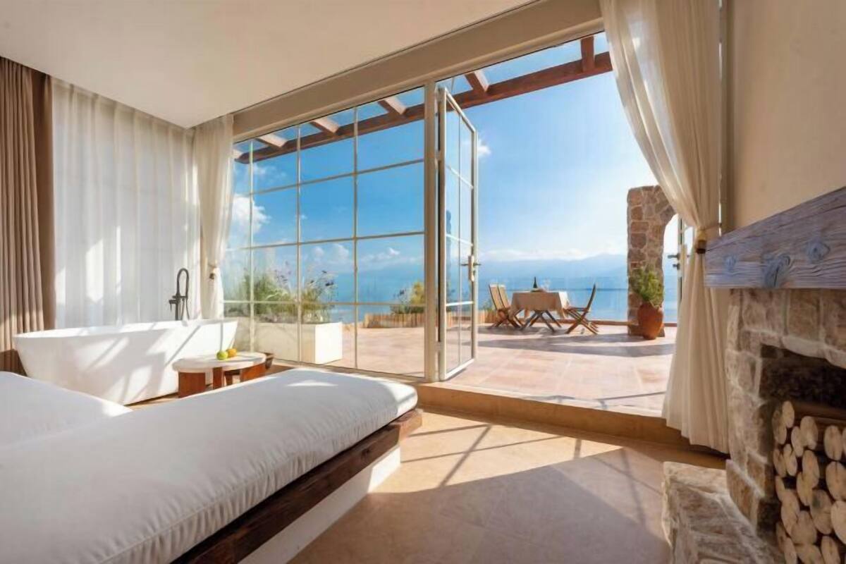 A bedroom with a large bed, a bathtub, and a view of the sea through a glass door.