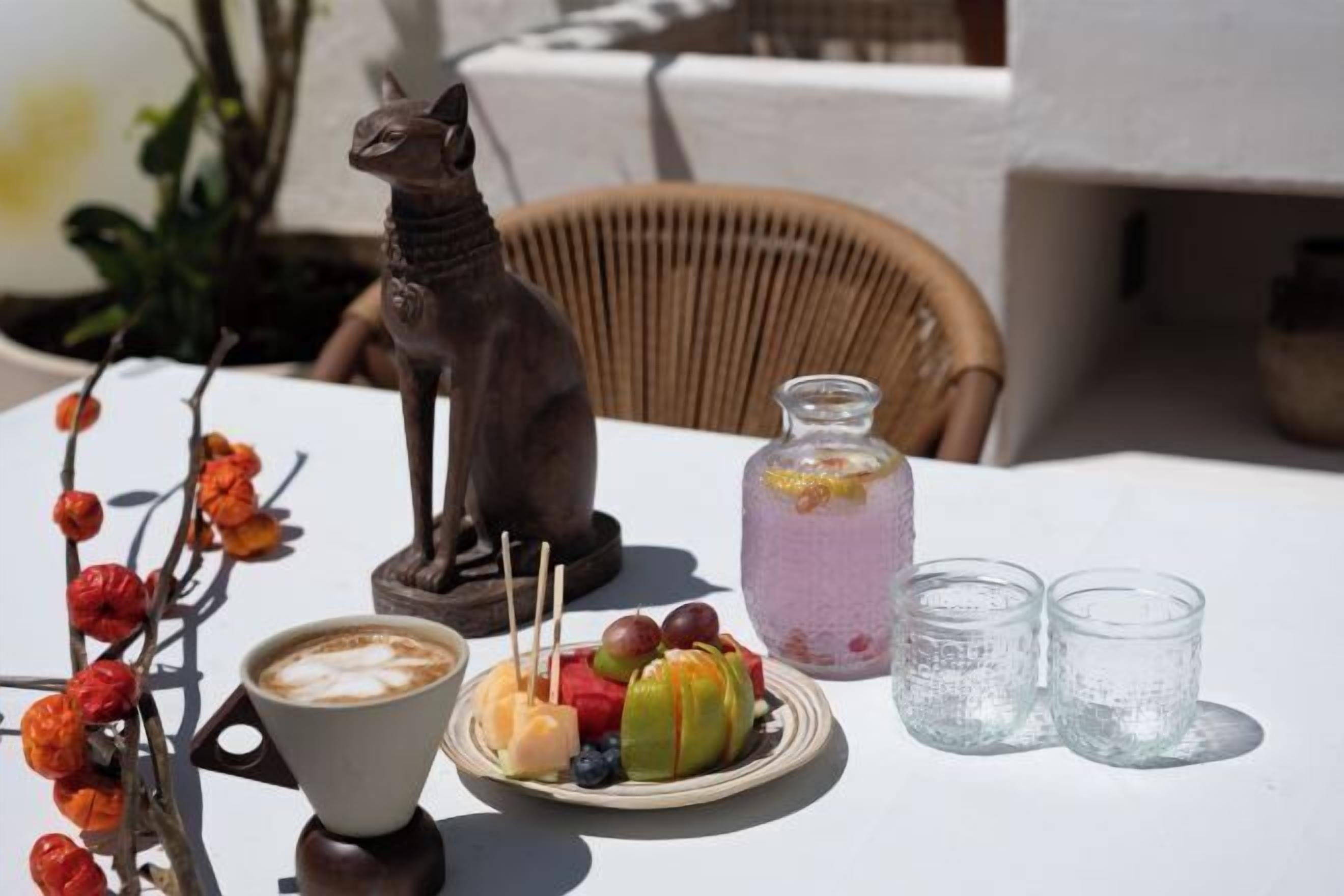 A table with a coffee cup, fruit, a decorative cat statue, a glass of pink drink, and two clear glasses.