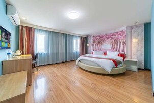 Quarto de hotel com cama redonda, escrivaninha, cadeira e uma janela grande com cortinas.