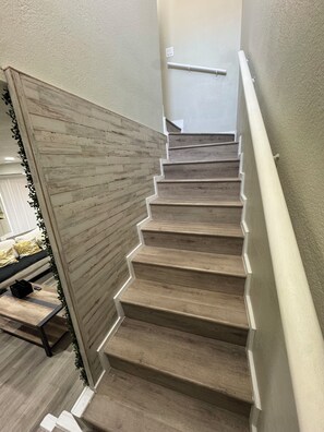 Un escalier avec des marches en bois et une rampe blanche, menant à une pièce où se trouvent un lit et une table de chevet.