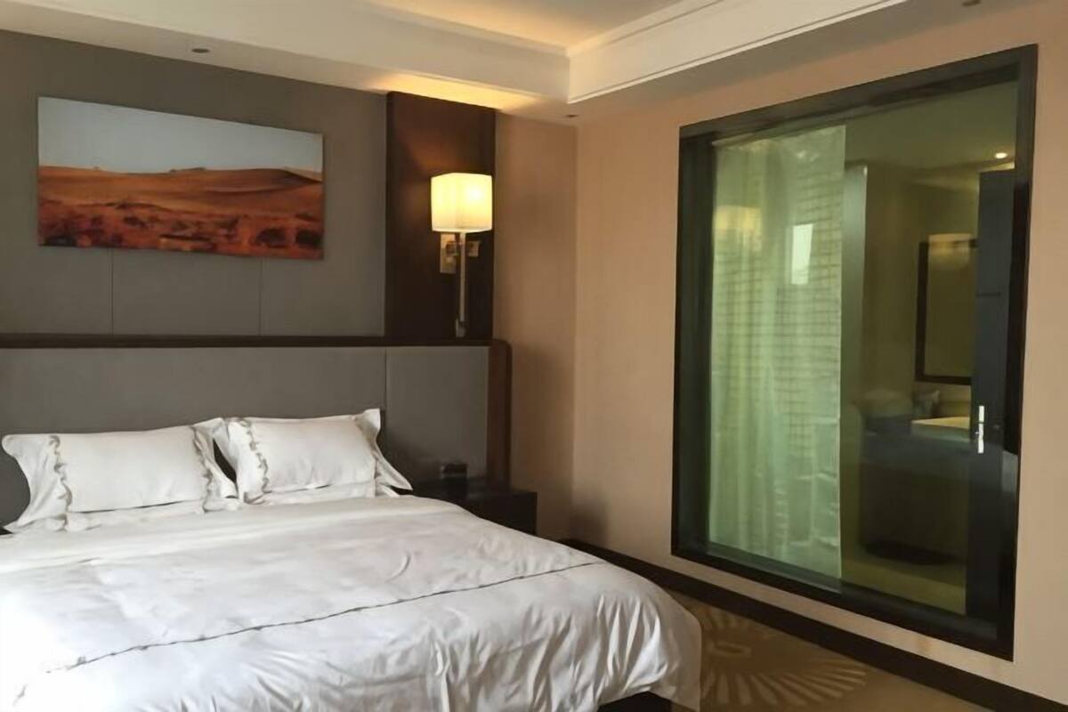 A hotel room with a large bed, a nightstand, a wall-mounted lamp, a painting, and a large window with a view.