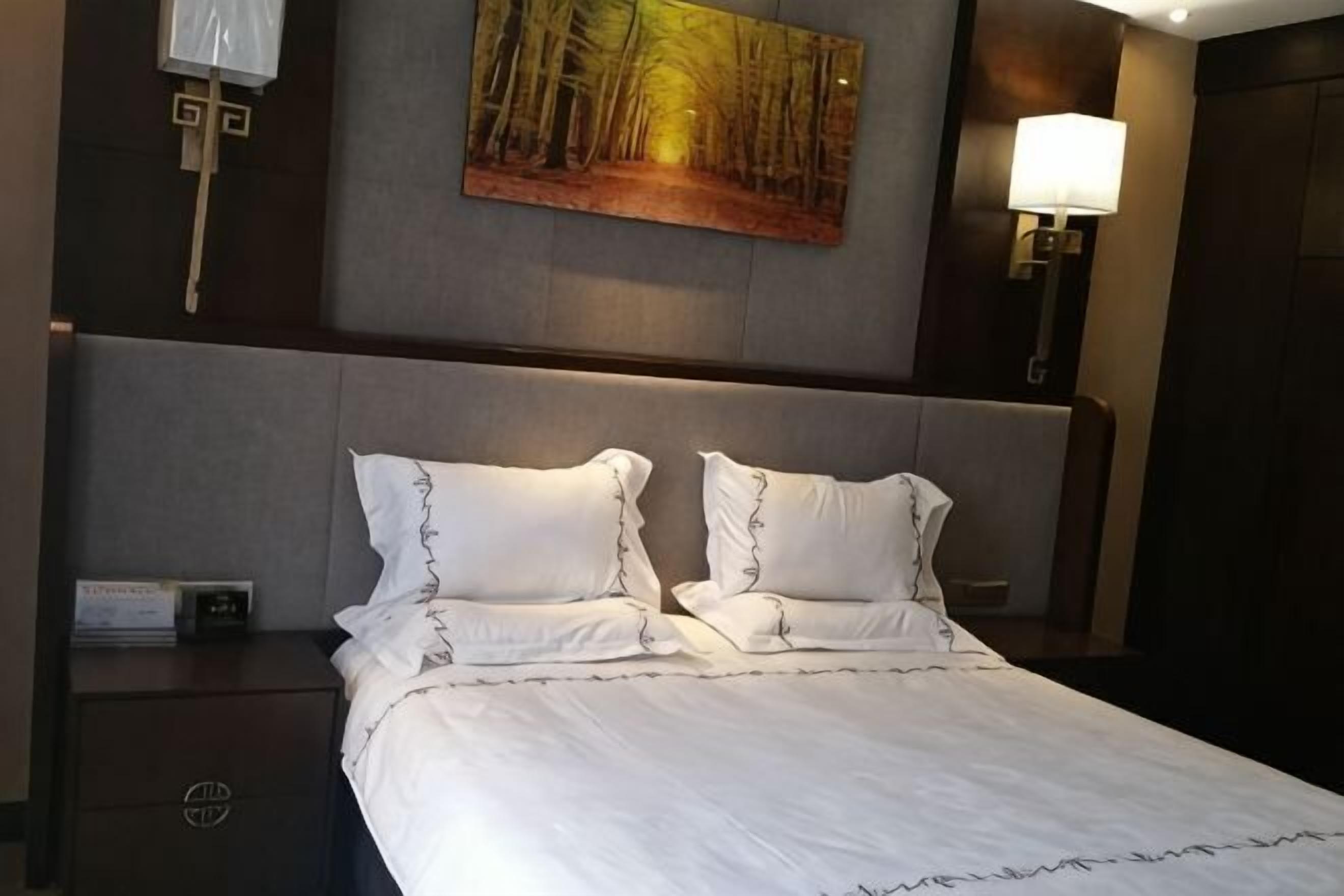 A hotel room with a large bed, a nightstand, a wall-mounted lamp, and a painting above the bed.