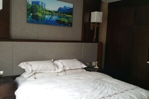 A neatly made bed with white linens and decorative pillows, a painting of a landscape on the wall, and a bedside lamp. - KAIYUE HOTEL (Qiemo)
