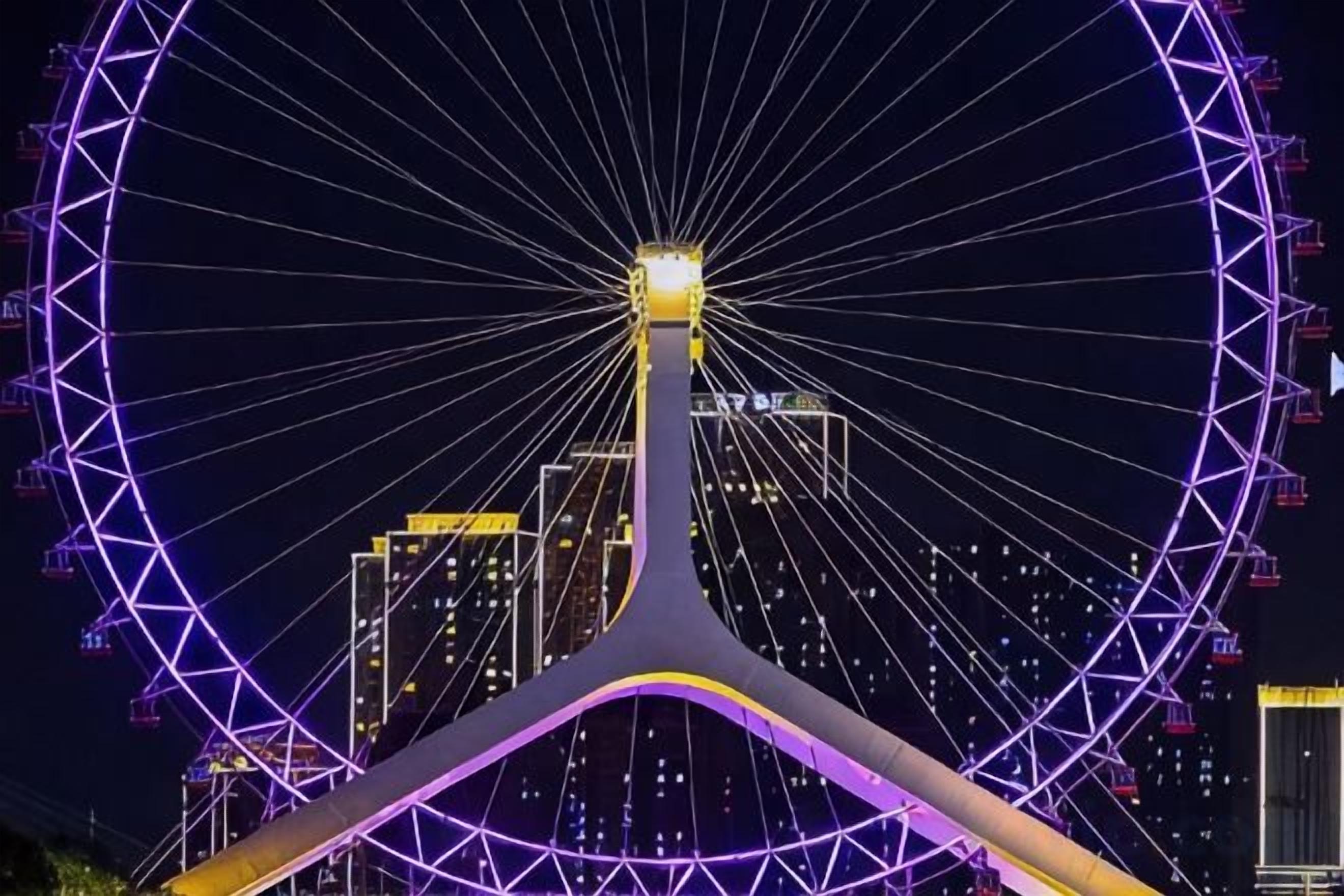 A large Ferris wheel with a central hub and multiple spokes, surrounded by a cityscape at night.
