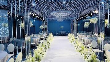A venue with a central aisle leading to a stage, surrounded by round tables and chairs, with a ceiling structure and floral decorations.