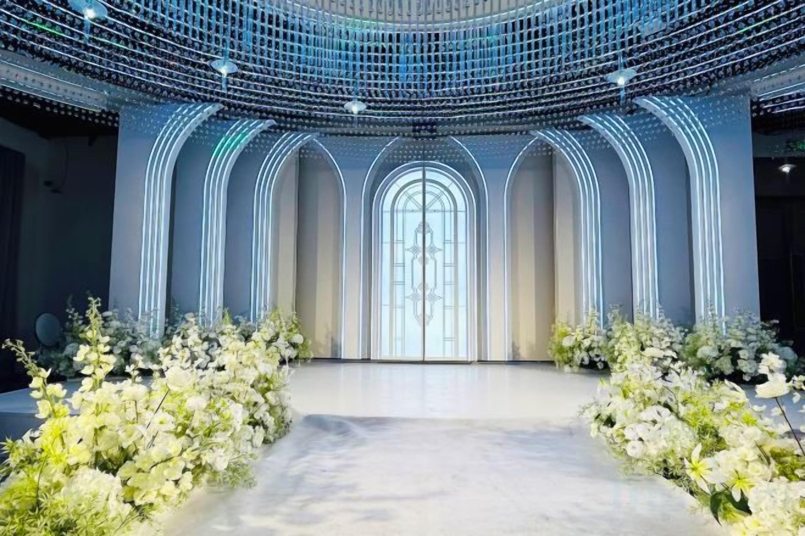 Un espacio con un pasillo central bordeado por arreglos florales blancos, que conduce a un amplio arco de entrada.