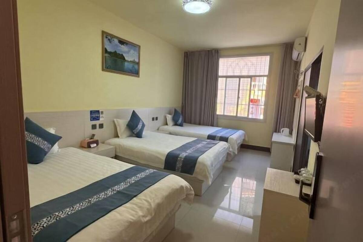 A hotel room with three beds, a window with curtains, a painting on the wall, and a television on the right.