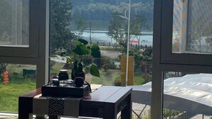 A room with a large window offering a view of a lake and surrounding greenery. There is a small table with a potted plant and a decorative box.