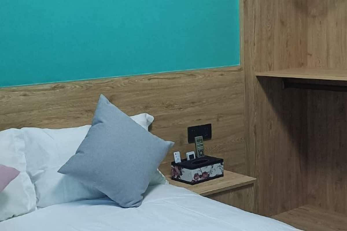 A neatly made bed with white and light blue pillows, a wooden headboard, and a nightstand with a tissue box and a phone charger.