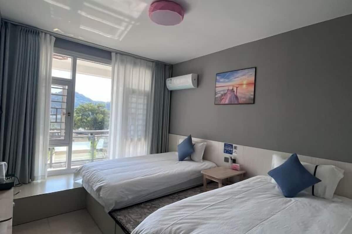 A hotel room with two beds, a small table, an air conditioning unit, a painting, and a view through the window.