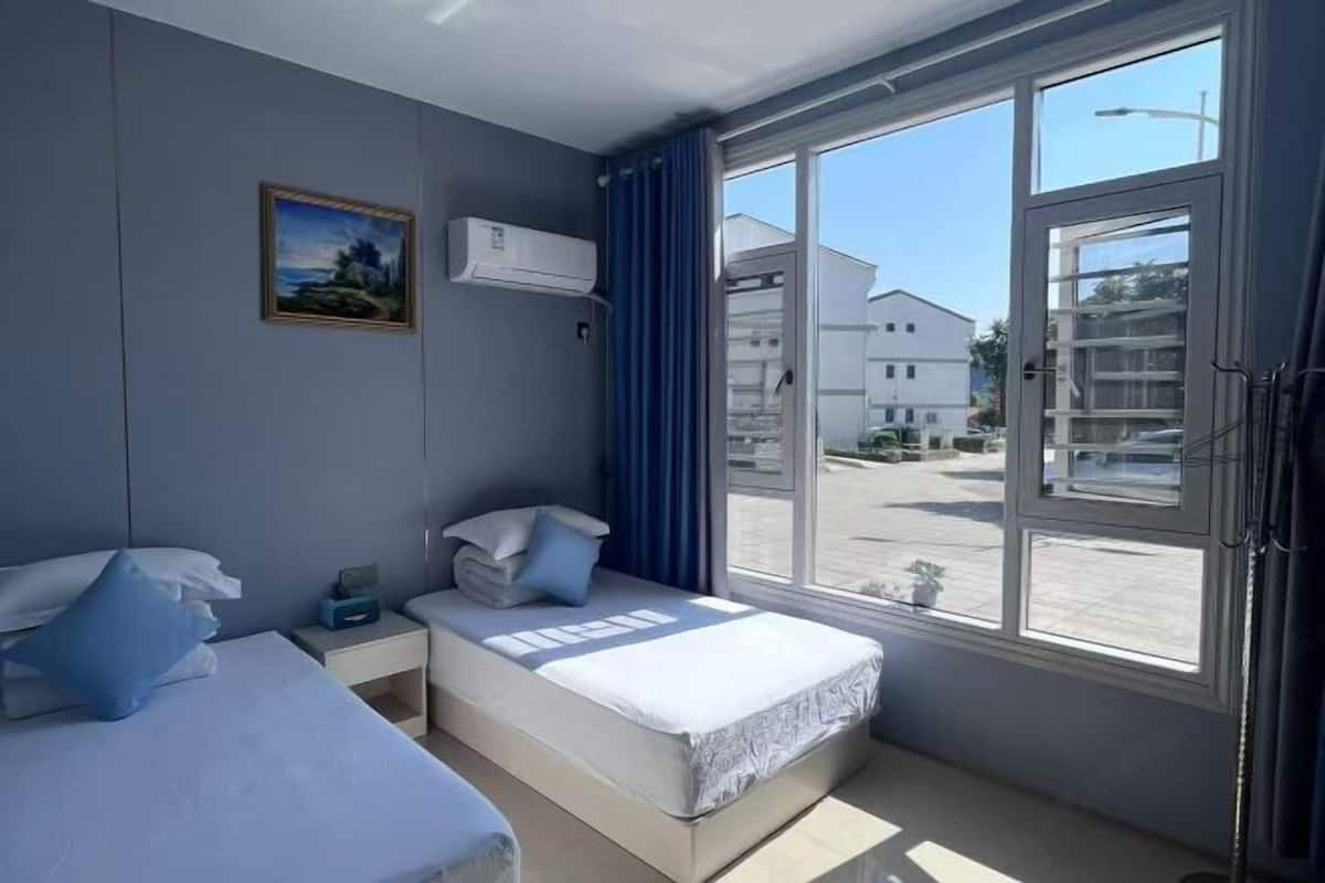 A bedroom with a bed, a nightstand, an air conditioning unit, a window with curtains, and a view of the outside.