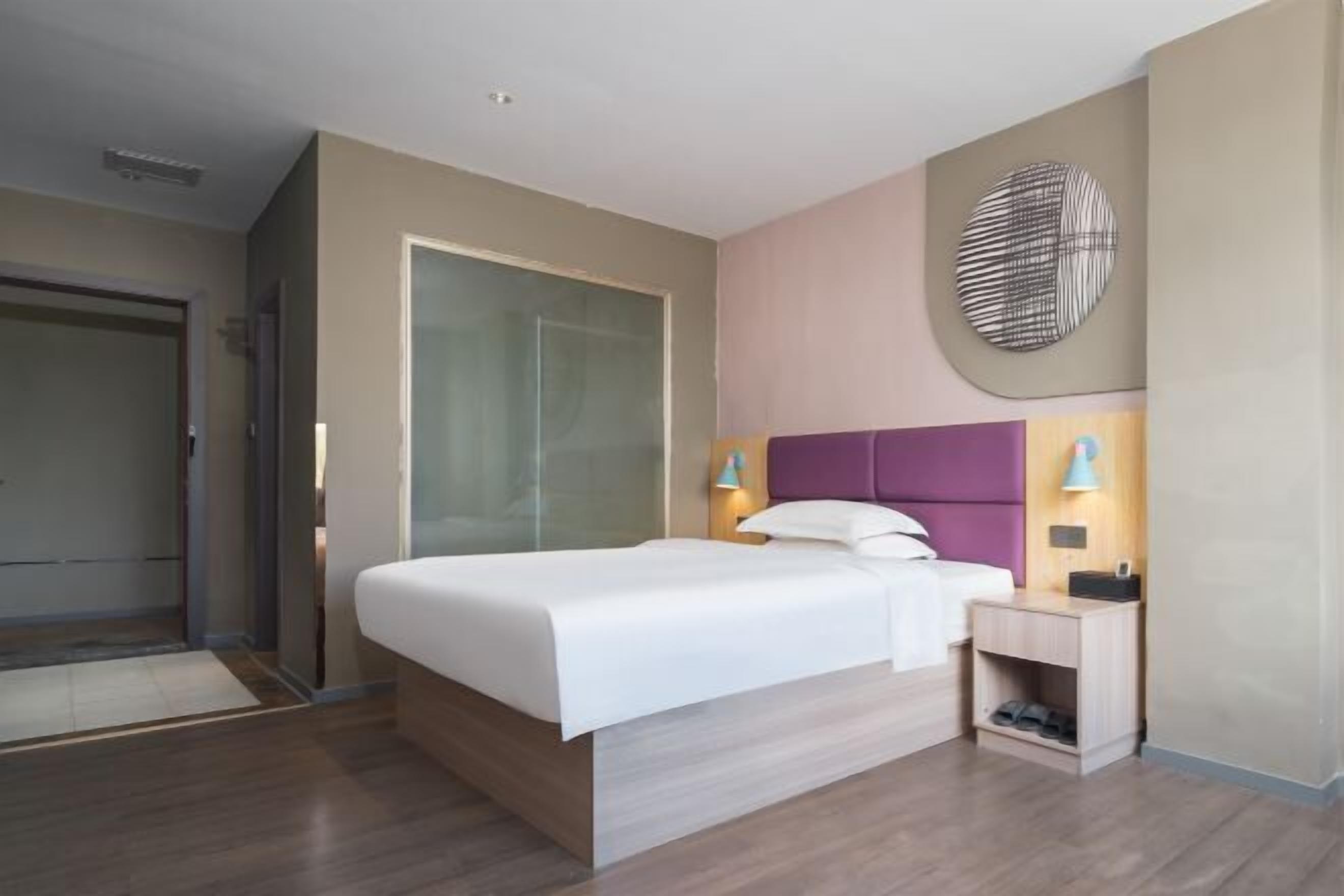 A modern hotel room with a large bed, a nightstand, a wall-mounted lamp, and a door to the left.