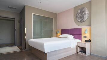 A modern hotel room with a large bed, a nightstand, a wall-mounted lamp, and a door to the left.