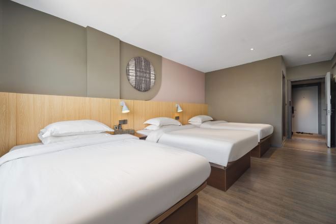 A hotel room with three beds, wooden headboard, wall-mounted lamps, and a circular wall decoration.
