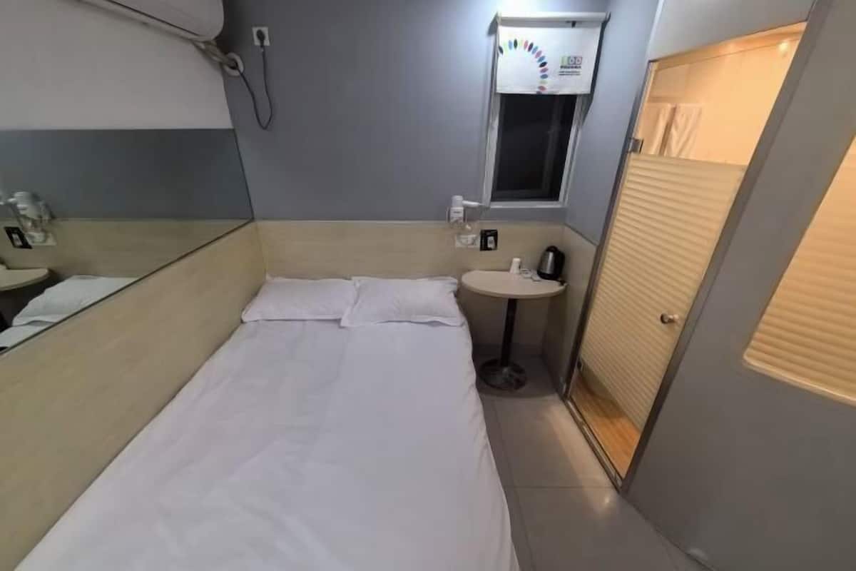 A compact hotel room with a single bed, a small bedside table, a window with a curtain, and a mirror.