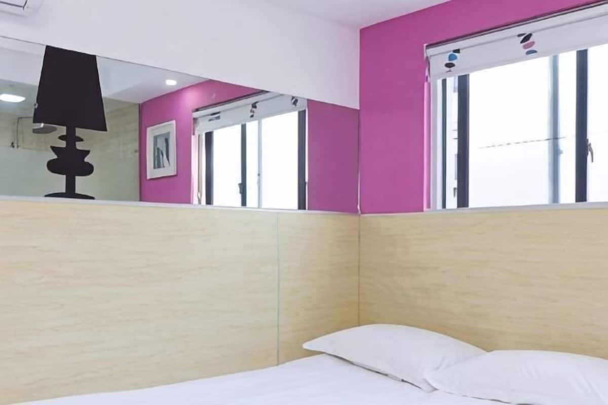 A room with a bed, a mirror, a window with blinds, and a pink wall.