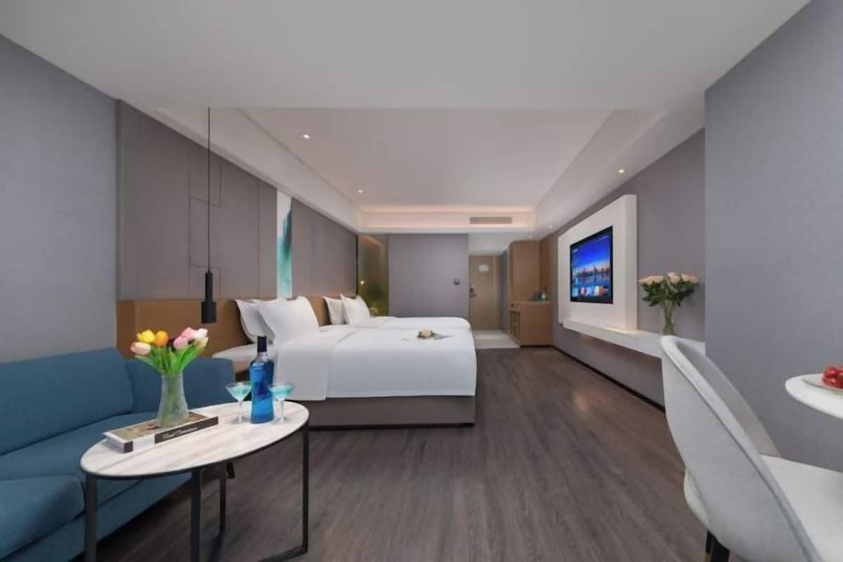 A modern hotel room with a bed, a sofa, a small table with a vase of flowers, and a TV on the wall.