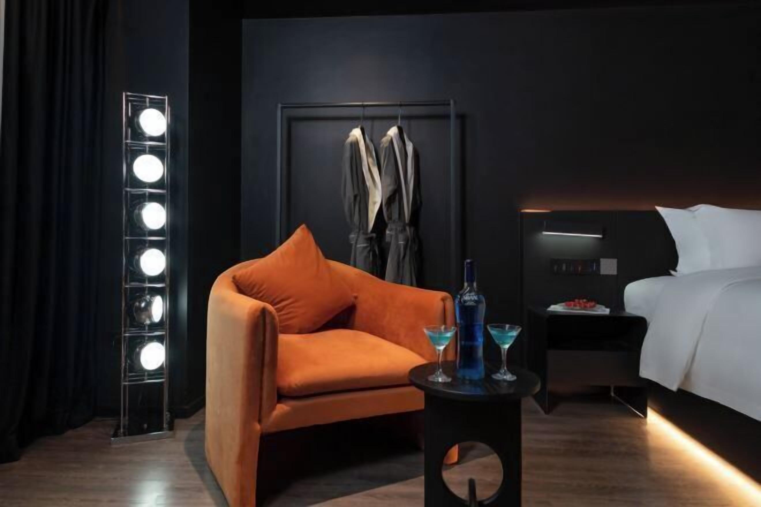A modern hotel room with a curved orange armchair, a bedside table with two bottles, and a headboard with a light strip.