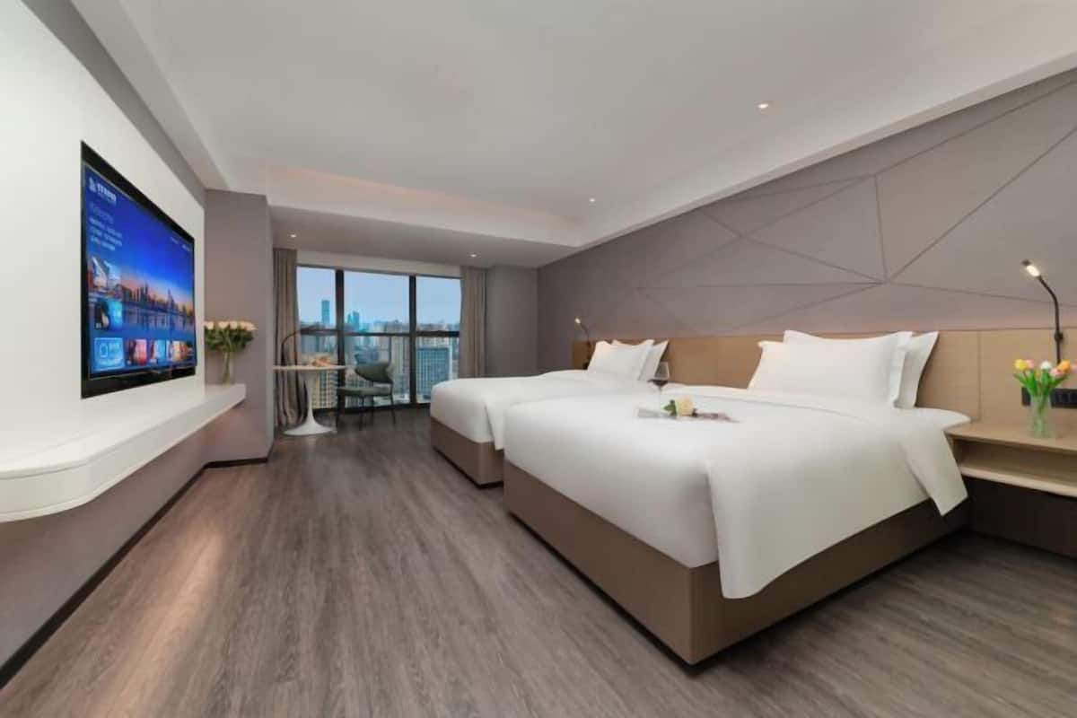 A modern hotel room with two beds, a large TV on the wall, and a city view.
