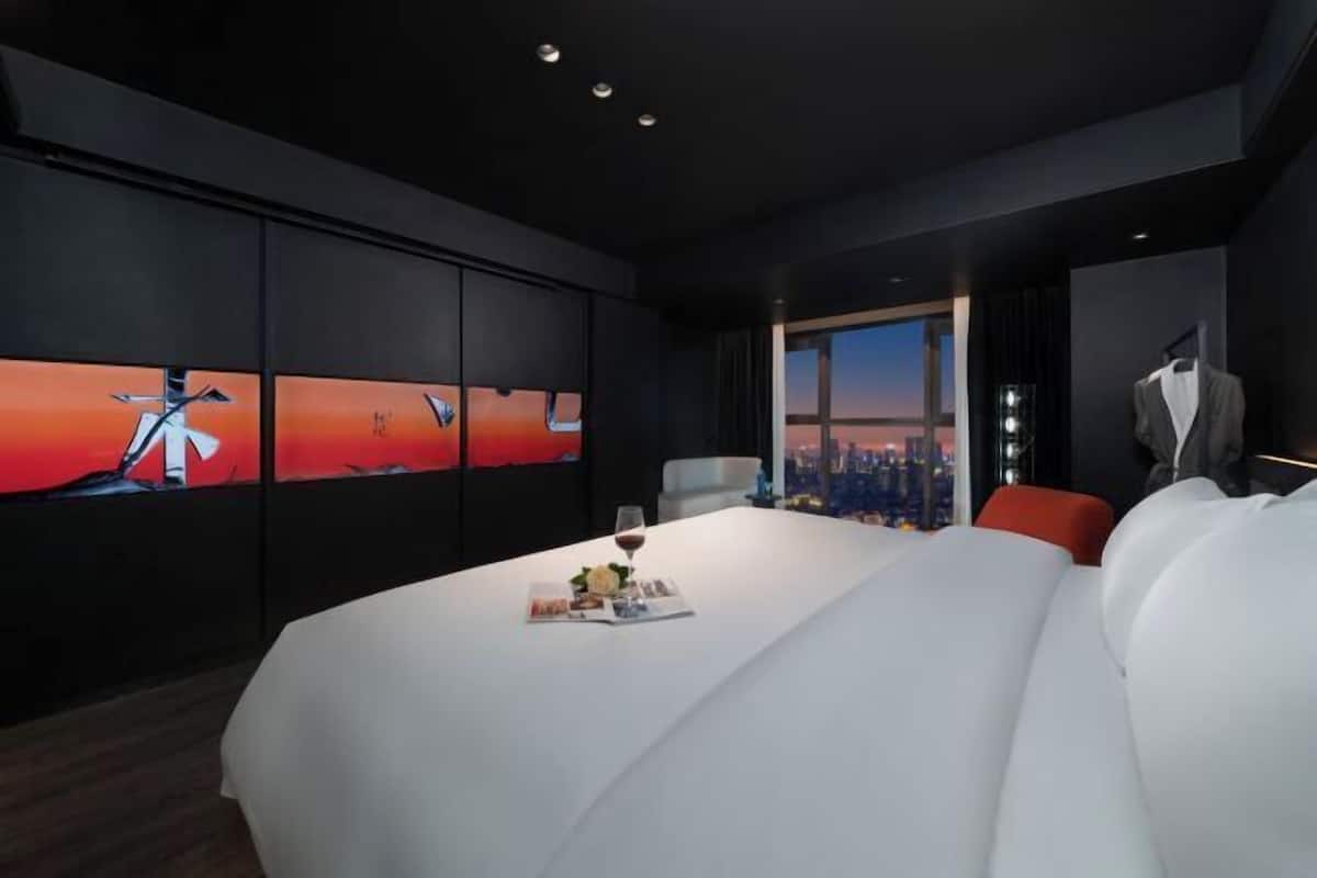 A modern hotel room with a large bed, a dining table with a wine glass, and a cityscape view through the window.