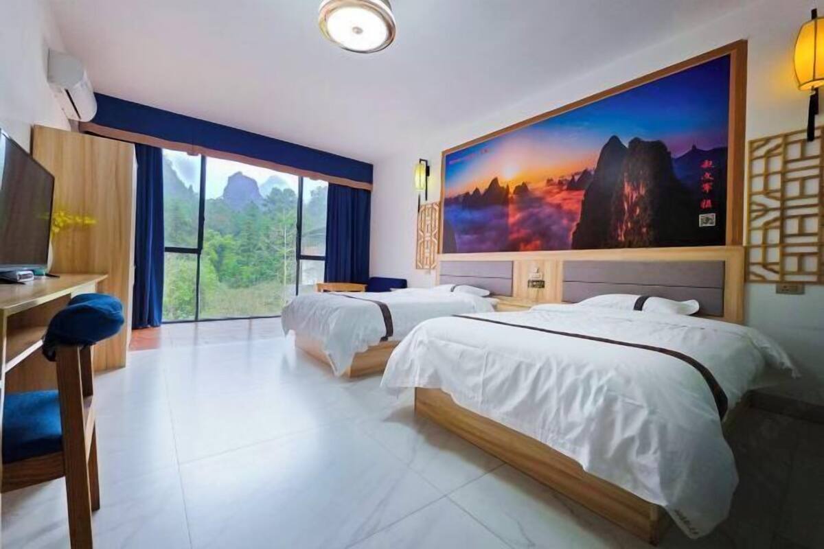 Habitación de hotel con dos camas, un escritorio, televisión y una ventana grande con vistas a la montaña.