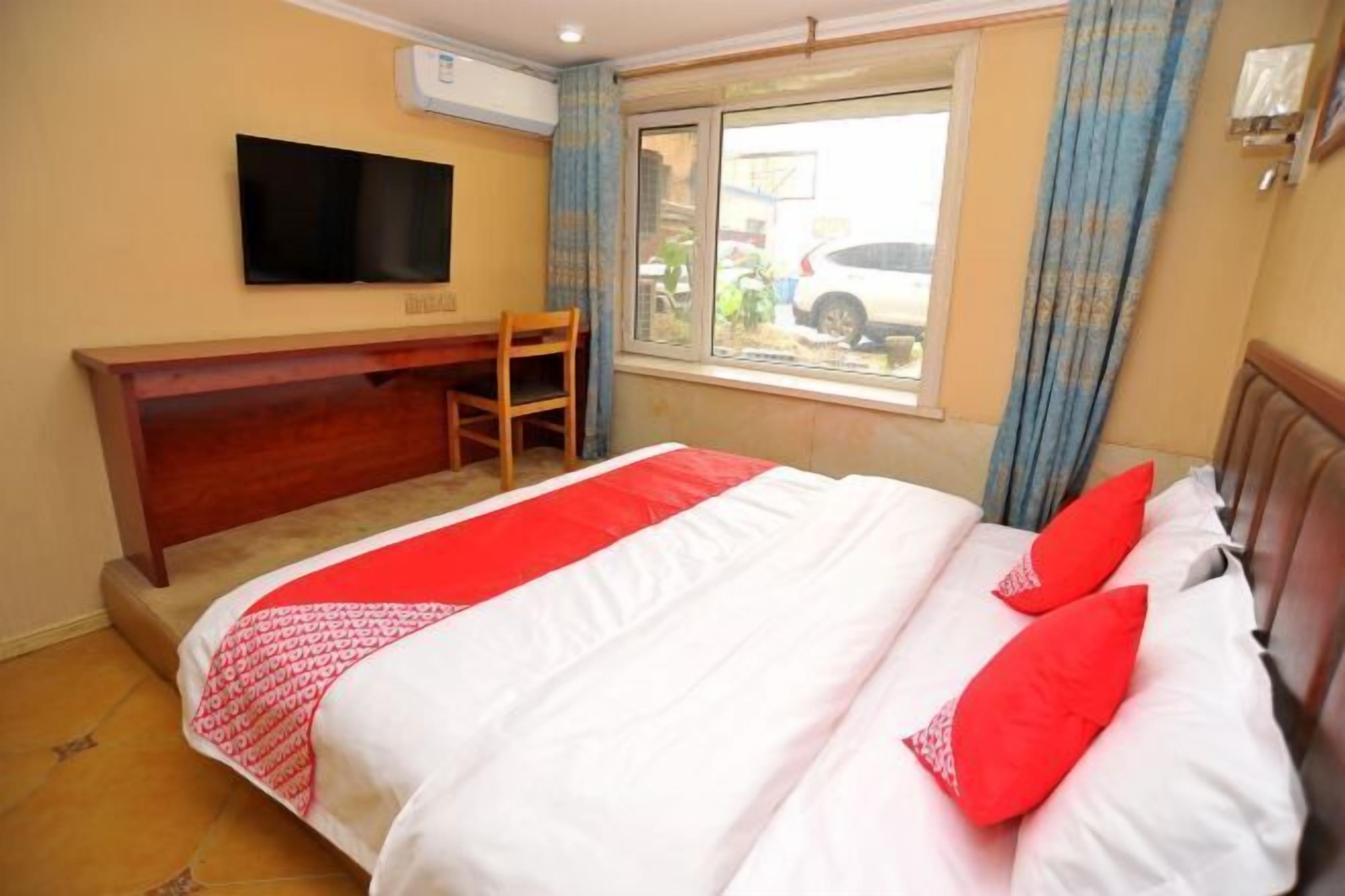 A hotel room with a bed, a TV mounted on the wall, a window with curtains, and a wooden desk.