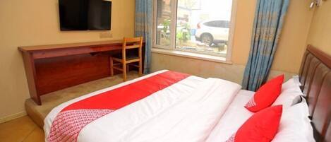A hotel room with a bed, a TV mounted on the wall, a window with curtains, and a wooden desk.
